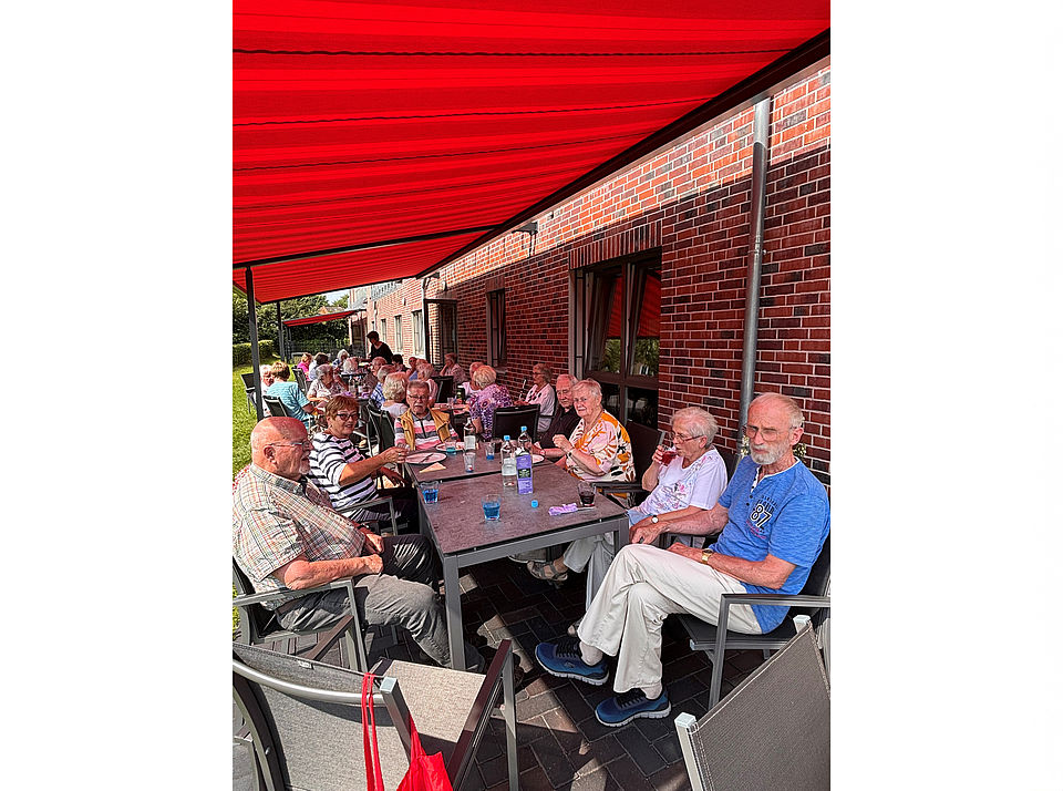 Eine große Gruppe Senioren sitzt fröhlich an Tischen auf der Terrasse unter der Markiese