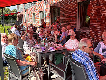 Eine fröhliche Gruppe SeniorInnen sitzt an Tischen auf der Terrasse unter der Markiese